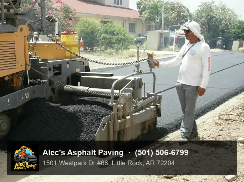 A worker at the screed controls of an asphalt paver as hot mix spreads across a residential street
