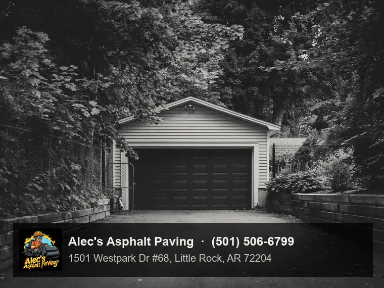 A dark asphalt driveway between timber retaining walls leading to a white detached garage with a black door, under heavy trees