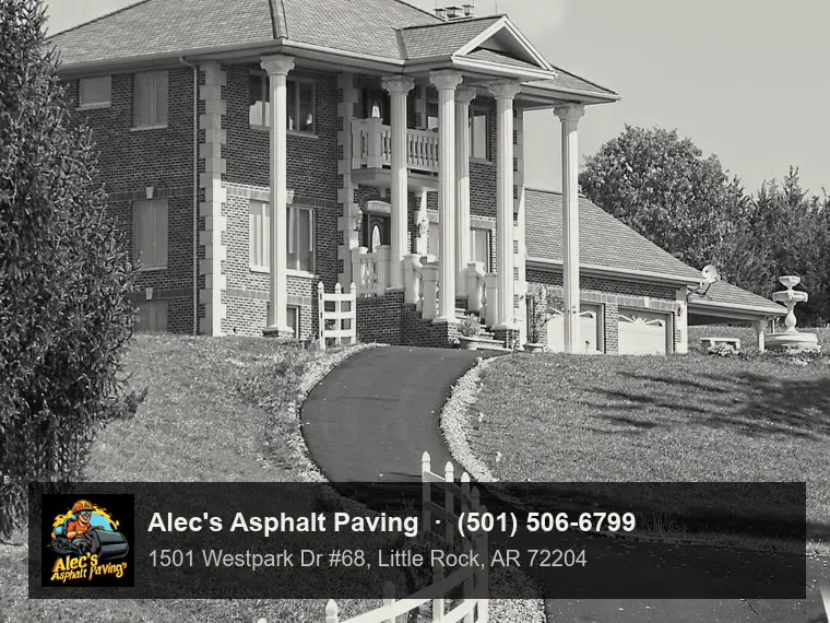 A long, deep-black asphalt driveway winding uphill past a white rail fence to a large brick house with tall white columns