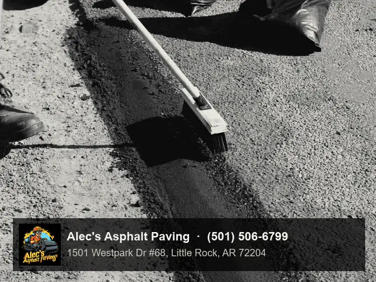 A worker brushing a narrow strip of fresh black asphalt patching laid along the edge of a stony road, a bag of patch material behind