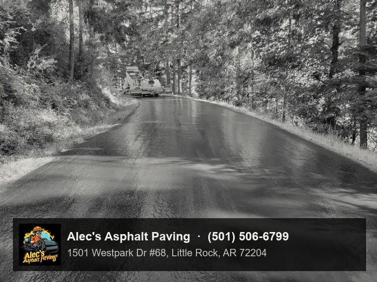 A freshly sprayed, still-glossy road surface curving through woods, with a dump truck and chip spreader in the distance