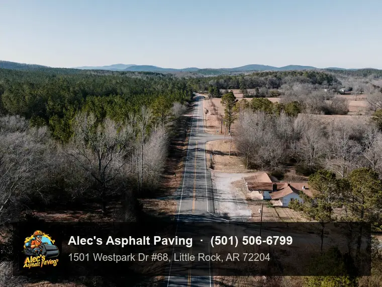 Drone view along a two-lane highway running through pine forest and winter-bare hardwoods toward low forested ridges, a farmhouse and side road on the right