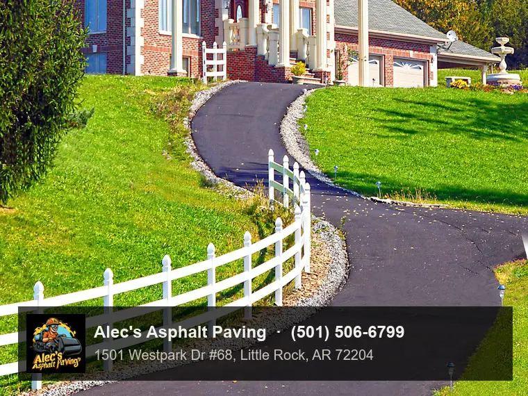 A long, deep-black asphalt driveway winding uphill past a white rail fence and solar path lights to a large brick house with tall columns, on a sunny day