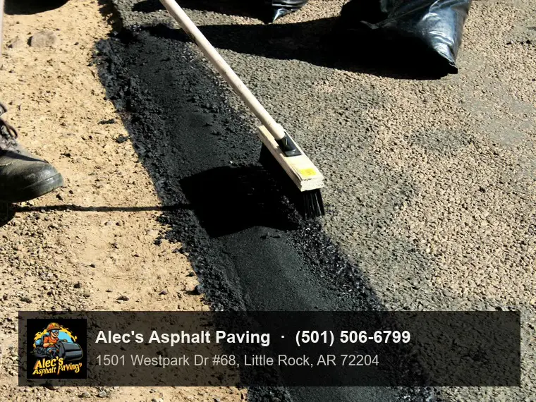 A worker brushing the surface of a narrow strip of fresh asphalt patching laid along the edge of a road beside bare dirt, bags of patch material behind