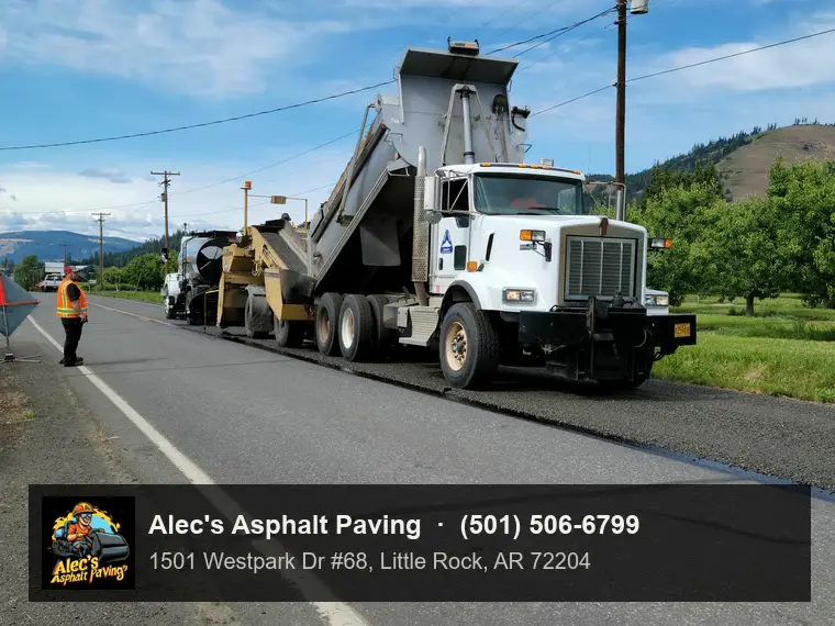 A dump truck backing into a chip spreader on a two-lane road during a chip-seal job