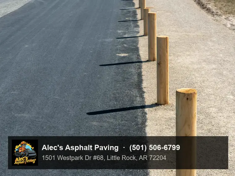 A freshly paved, smooth black asphalt lane with a clean edge against a sandy gravel shoulder lined with round wooden posts, in bright sun