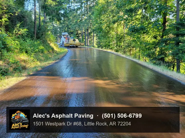 A freshly sprayed, still-glossy road curving through woods, with a chip spreader and dump truck working in the distance