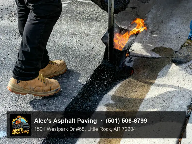 Close view of a worker's boots beside a hand pour pot laying a band of hot, gritty sealant along the seam where asphalt meets a concrete gutter, a flame flaring at the pot