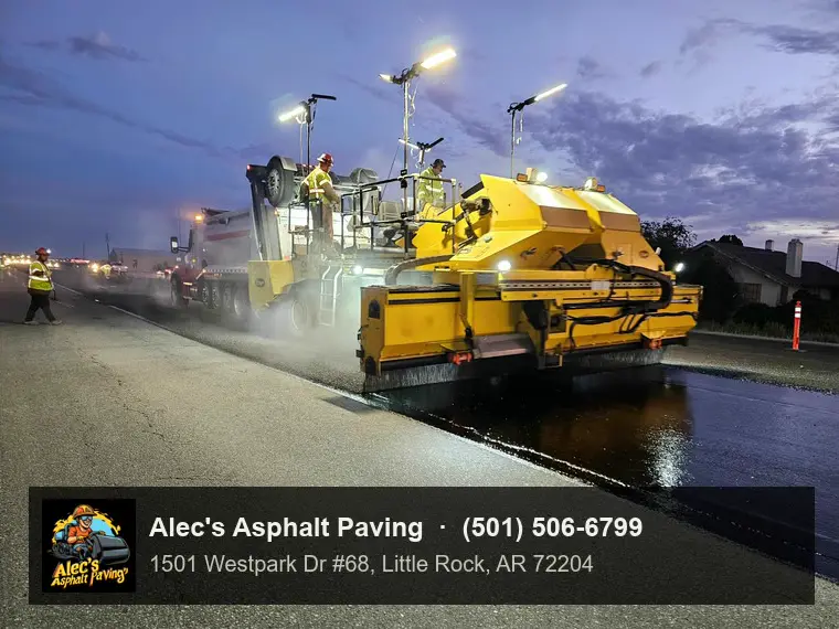 A chip spreader working under its own lights at dusk over freshly sprayed asphalt, a crew member walking alongside